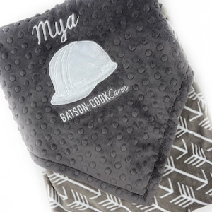 May include: A gray and white baby blanket with a white embroidered design of a hard hat and the text "Mya BATSON-COOK Cares".