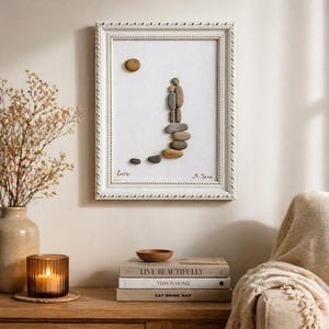 May include: A framed pebble art piece with a white frame. The art depicts a couple made of pebbles walking up a path of stones. The word "love" is written at the bottom left. A sun-shaped pebble is at the top. Signed "N. Jane".
