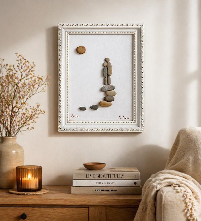 May include: A framed pebble art piece with a white frame. The art depicts a couple made of pebbles walking up a path of stones. The word "love" is written at the bottom left. A sun-shaped pebble is at the top. Signed "N. Jane".