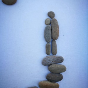May include: A minimalist artwork featuring a family silhouette and a sun, all created with smooth, rounded stones. The stones vary in size and color, arranged against a light blue background. The artwork evokes a sense of peace and simplicity.