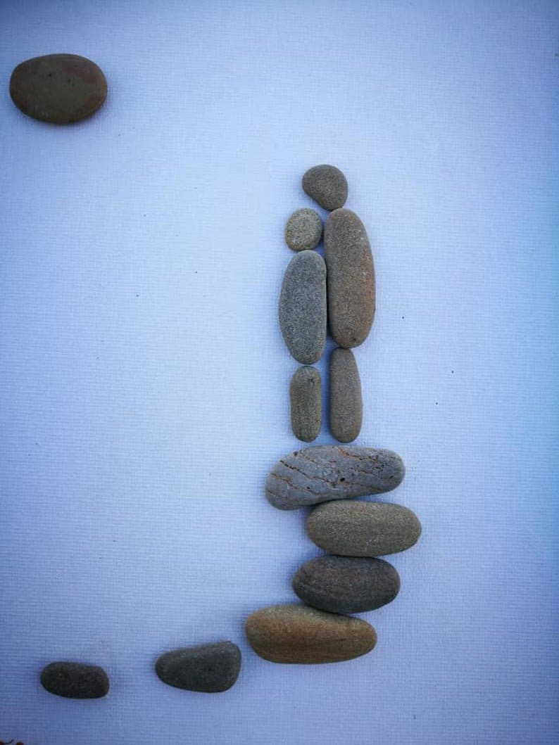 May include: A minimalist artwork featuring a family silhouette and a sun, all created with smooth, rounded stones. The stones vary in size and color, arranged against a light blue background. The artwork evokes a sense of peace and simplicity.