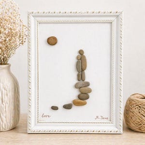 May include: Framed pebble art depicting a family. The artwork features a white frame with a textured design. The image shows a sun and a family made of pebbles. The word "love" is written in cursive. Signed "M. Yane".