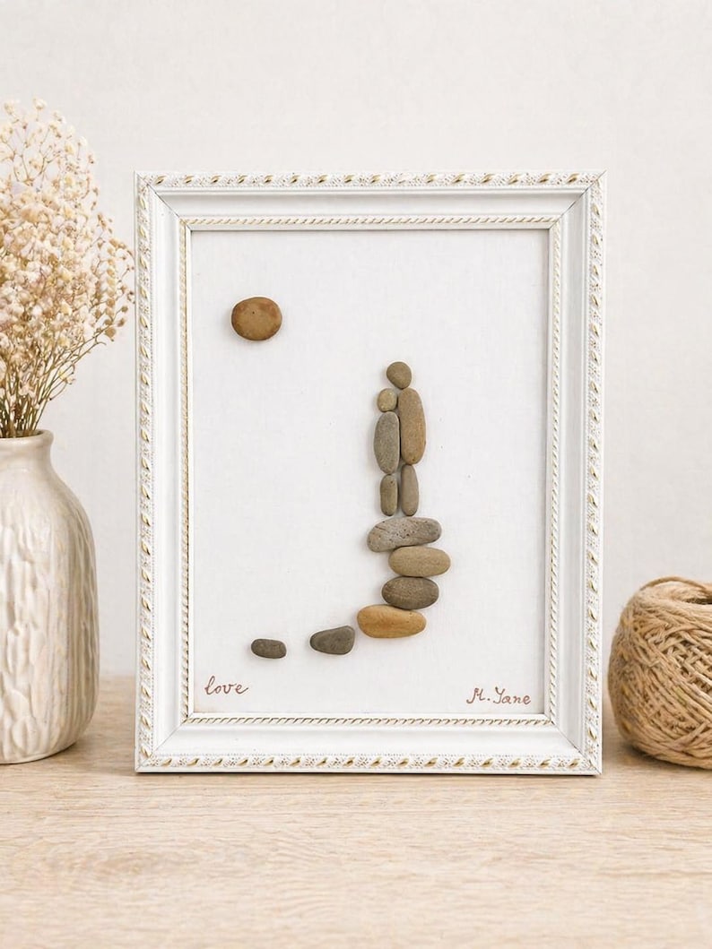 May include: Framed pebble art depicting a family. The artwork features a white frame with a textured design. The image shows a sun and a family made of pebbles. The word "love" is written in cursive. Signed "M. Yane".