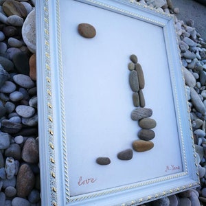 May include: Framed pebble art depicting a couple standing on a stack of stones, with a large pebble representing the sun. The white frame has a decorative gold trim. The word "love" is written in cursive.