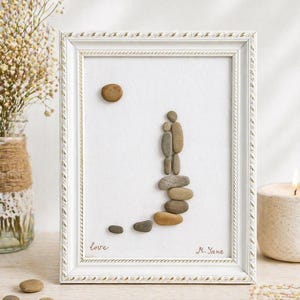 May include: A framed pebble art piece featuring a couple ascending a path. The artwork uses various sized pebbles to depict the figures and path, with a pebble sun. The frame is white with a decorative border, and the word "love" is written on the artwork.