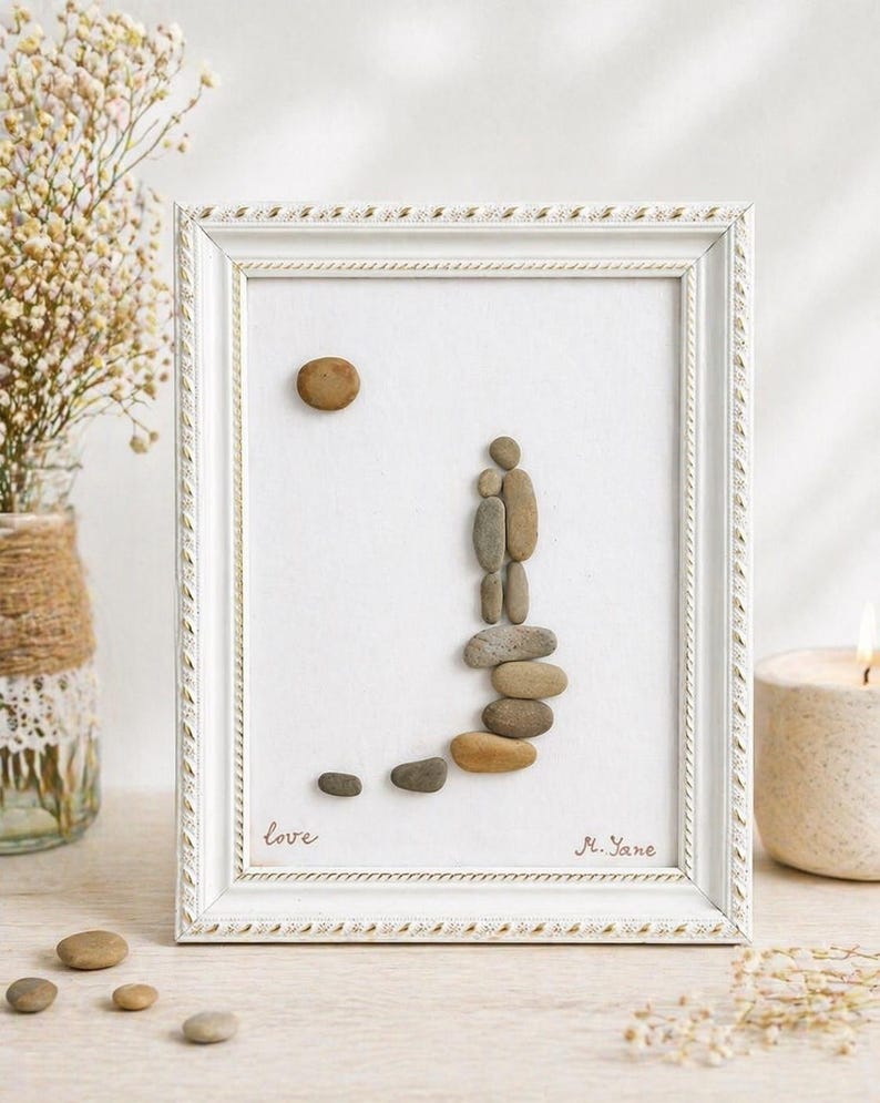 May include: A framed pebble art piece featuring a couple ascending a path. The artwork uses various sized pebbles to depict the figures and path, with a pebble sun. The frame is white with a decorative border, and the word "love" is written on the artwork.