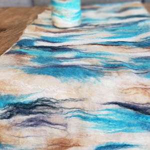以下が含まれることがあります： 青、茶色、白の波模様が施された手作りのフェルト製テーブルランナー。ランナーはウール製で、柔らかく質感のある表面をしています。