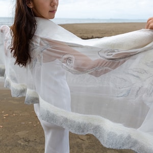May include: A white sheer shawl with a silver embroidered design. The shawl has a gray trim and is draped over a person's shoulders.
