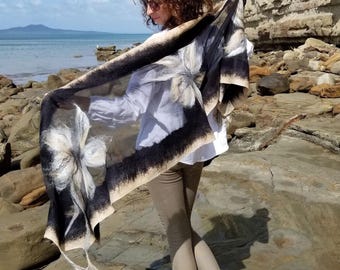 May include: A black and white floral scarf with a dark border is draped over the shoulders. The scarf features large, stylized flower designs. The person is wearing a white shirt, tan pants, and white sneakers. The background includes a rocky beach and ocean.