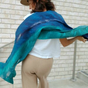 May include: A woman wearing a teal and blue ombre scarf with a white shirt and tan pants.