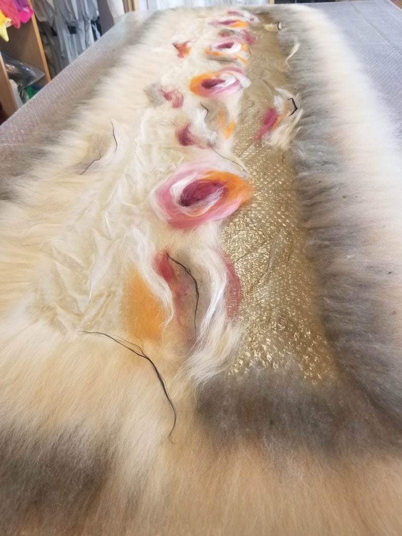 May include: A white, fluffy, felted fabric with a gold, sparkly, textured strip running down the center. The fabric is decorated with pink, orange, and white felt flowers.