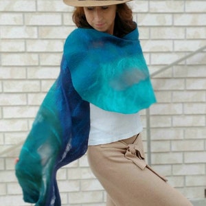 May include: A woman wearing a brown hat and a blue and green gradient scarf. The scarf is made of a soft, flowing fabric and is draped around her neck. She is wearing a white shirt and brown pants.