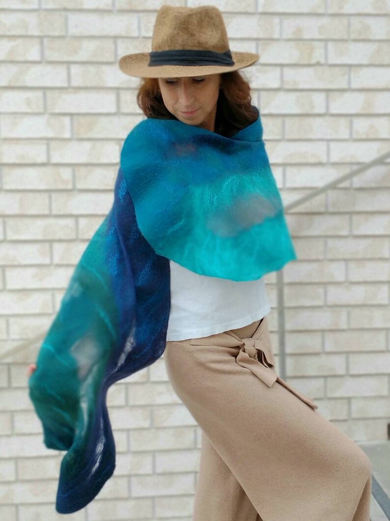 May include: A woman wearing a brown hat and a blue and green gradient scarf. The scarf is made of a soft, flowing fabric and is draped around her neck. She is wearing a white shirt and brown pants.