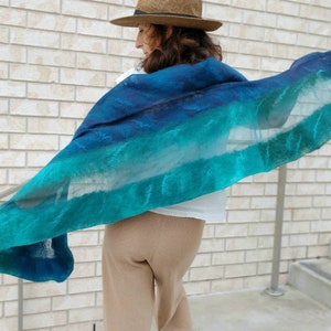 May include: A teal and blue ombre scarf with a sheer texture. The scarf is draped over a person's shoulders and is flowing in the wind.