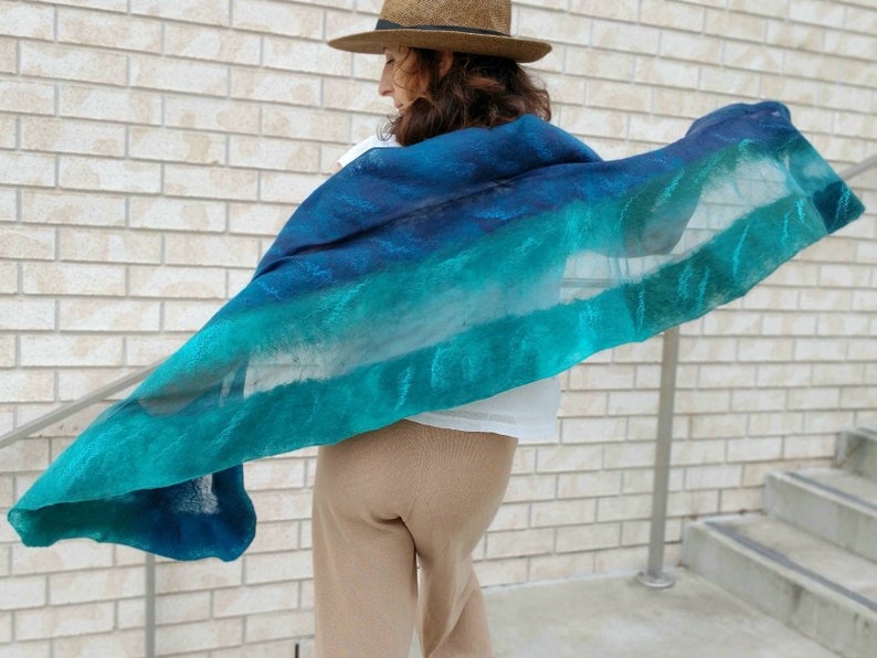 May include: A teal and blue ombre scarf with a sheer texture. The scarf is draped over a person's shoulders and is flowing in the wind.