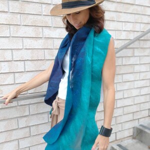 May include: A woman wearing a brown hat and a teal and blue gradient scarf. She is wearing a white sleeveless shirt and brown pants.