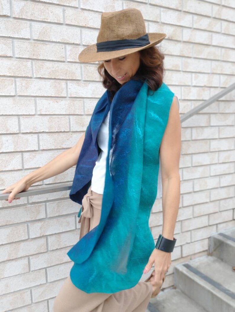 May include: A woman wearing a brown hat and a teal and blue gradient scarf. She is wearing a white sleeveless shirt and brown pants.