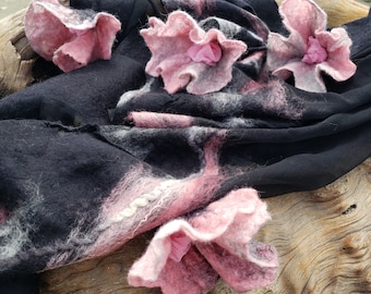 Könnte beinhalten: Ein schwarzer und rosa Wollschal mit angehängten rosa Filzblumen. Der Schal liegt über einem Stück Treibholz.