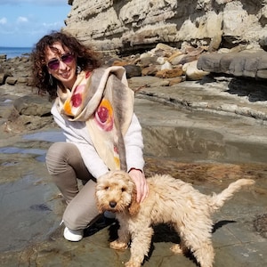 May include: A woman wearing sunglasses and a patterned scarf is kneeling on a rocky beach with a light brown dog. The dog is looking at the camera.