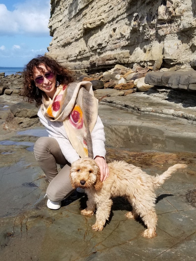 May include: A woman wearing sunglasses and a patterned scarf is kneeling on a rocky beach with a light brown dog. The dog is looking at the camera.