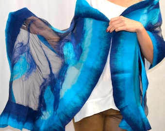 Può includere: Una sciarpa di seta blu e turchese con un design fluido. La sciarpa è drappeggiata sulle spalle di una persona ed è tenuta in posizione da una mano.