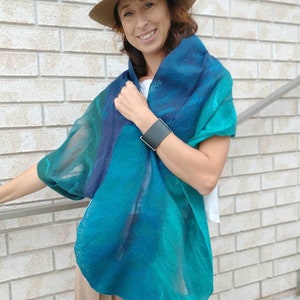 May include: A woman wearing a teal, blue, and green ombre scarf with a brown hat. She is wearing a white shirt and beige pants.