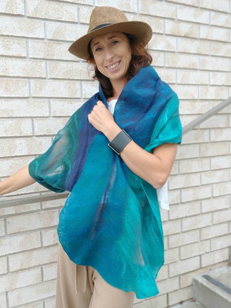 May include: A woman wearing a teal, blue, and green ombre scarf with a brown hat. She is wearing a white shirt and beige pants.