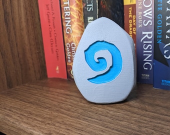 Könnte beinhalten: Ein graues, eiförmiges Dekorationsobjekt mit einem blauen Spiralmuster. Das Objekt steht auf einem Holzregal vor einer Reihe von Büchern mit farbigen Buchrücken. Ein einzigartiges Dekorationsobjekt.