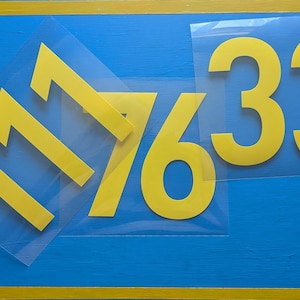 Könnte beinhalten: Gelbe Klebezahlen 1, 7, 6 und 3 auf blauem Hintergrund. Die Zahlen sind in einer fetten, serifenlosen Schriftart gehalten. Der Hintergrund ist eine lackierte blaue Oberfläche mit gelbem Rand. Die Zahlen befinden sich auf einer transparenten Folie.