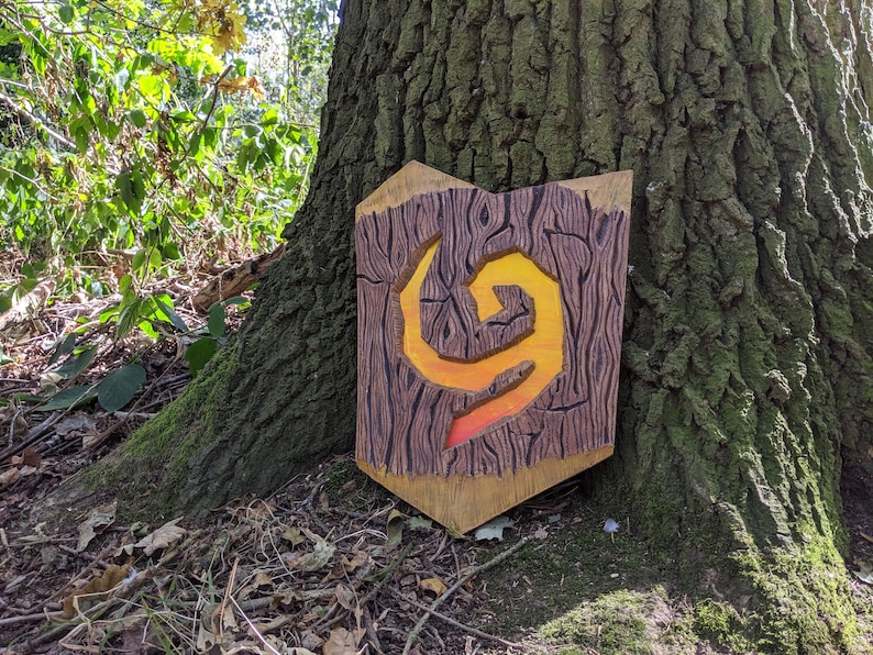 Puede incluir: Un escudo de madera con un dise&ntilde;o tallado de una llama giratoria en tonos naranja y amarillo. El escudo est&aacute; apoyado contra un tronco de &aacute;rbol.