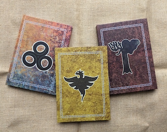 Puede incluir: Tres cuadernos diferentes con diseños únicos. El primer cuaderno tiene un fondo azul, naranja y marrón con un símbolo negro de tres círculos. El segundo cuaderno tiene un fondo dorado con un símbolo de pájaro negro. El tercer cuaderno tiene un fondo marrón con un símbolo de árbol negro.