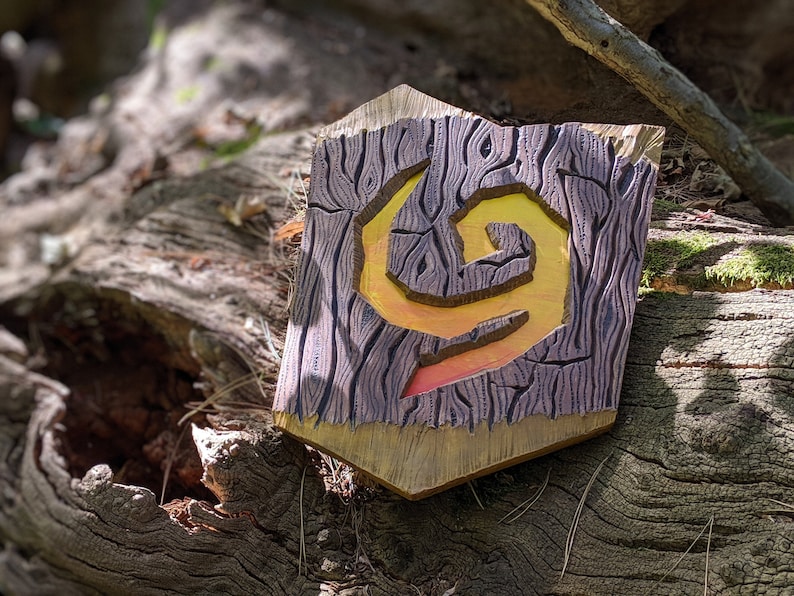 Puede incluir: Un escudo de madera con un dise&ntilde;o tallado de una llama giratoria en tonos de amarillo, naranja y rojo. El escudo est&aacute; pintado para que parezca veta de madera.