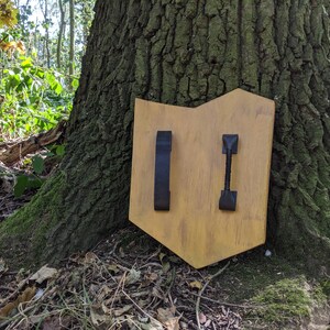 Puede incluir: Un escudo de madera con dos correas de cuero negro. El escudo est&aacute; pintado de color marr&oacute;n claro y tiene un aspecto ligeramente desgastado.