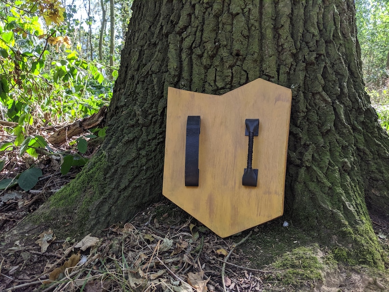 Puede incluir: Un escudo de madera con dos correas de cuero negro. El escudo est&aacute; pintado de color marr&oacute;n claro y tiene un aspecto ligeramente desgastado.