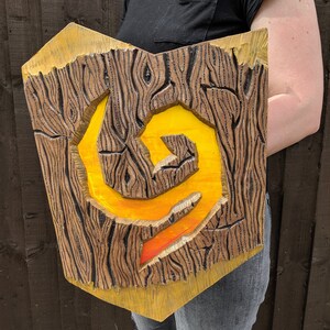 Puede incluir: Un escudo de madera con un s&iacute;mbolo amarillo y naranja pintado en &eacute;l. El escudo est&aacute; tallado para parecer madera y tiene un borde elevado.