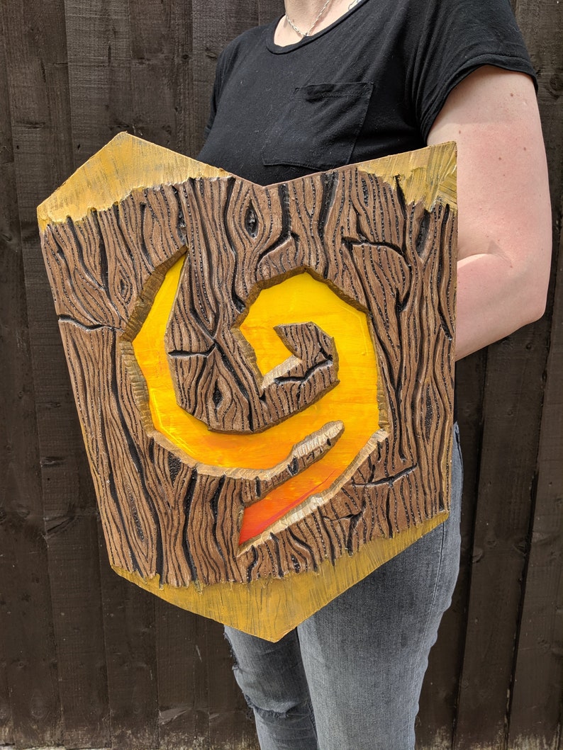 Puede incluir: Un escudo de madera con un s&iacute;mbolo amarillo y naranja pintado en &eacute;l. El escudo est&aacute; tallado para parecer madera y tiene un borde elevado.