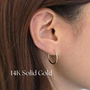 May include: A close-up of a person's ear with a small, gold hoop earring. The earring is made of 14K solid gold.