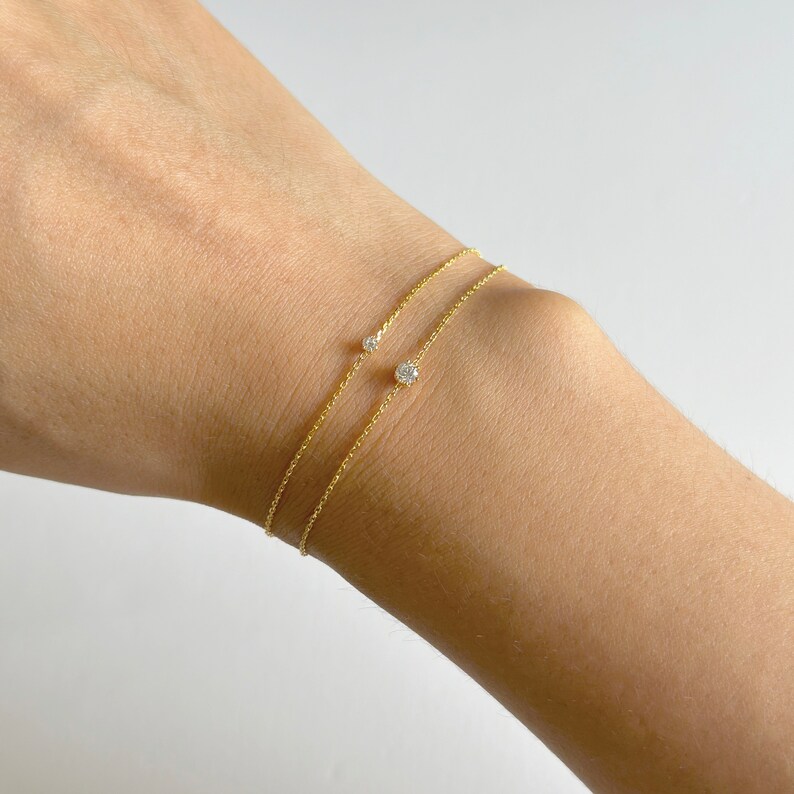 May include: Two delicate gold chain bracelets with small diamond accents. The bracelets are layered on a person's wrist.
