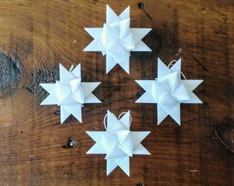 May include: Four white paper star ornaments with pointed edges and a loop for hanging, arranged on a rustic wooden surface. The stars have a geometric, folded design, suggesting a handcrafted or DIY aesthetic, perfect for holiday decor.