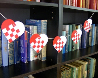 May include: A red and white checkered heart garland hangs on a bookshelf filled with books. The garland is made of paper and is strung together with twine.