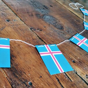 May include: A string of Icelandic flags, each featuring a white cross on a blue background with a pink stripe, hangs on a wooden surface.