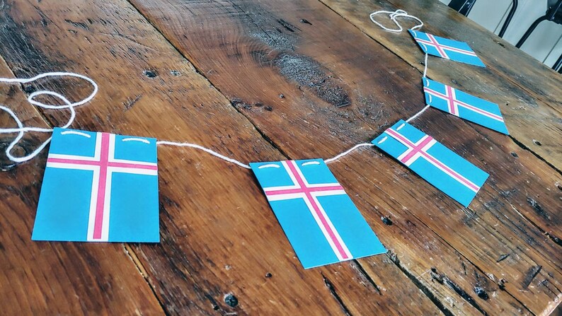 May include: A string of Icelandic flags, each featuring a white cross on a blue background with a pink stripe, hangs on a wooden surface.