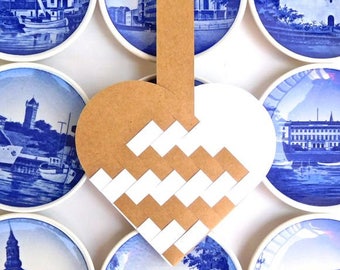 Pode incluir: Uma coleção de pratos decorativos azuis e brancos com várias cenas de paisagens. Um coração artesanal feito de papel branco e castanho tecido é centrado sobre os pratos, criando uma composição visual única. Os pratos estão dispostos sobre uma superfície branca.