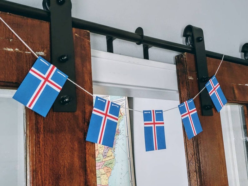 May include: A string of Icelandic flags hanging from a black metal track. The flags are blue with a white cross and a red stripe. The flags are hanging from a wooden door.