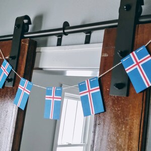 May include: A string of Icelandic flags hanging from a black metal sliding door track. The flags are blue with a white cross and red trim.