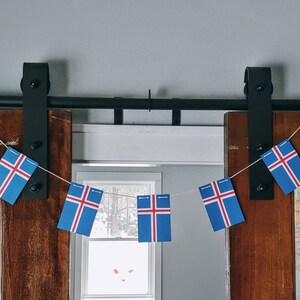 May include: A string of Icelandic flags hangs from a black metal track above a wooden door. The flags are blue, white, and red, and they are arranged in a row. The flags are hanging in front of a window with a view of a snowy landscape.