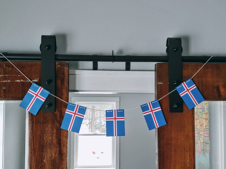 May include: A string of Icelandic flags hangs from a black metal track above a wooden door. The flags are blue, white, and red, and they are arranged in a row. The flags are hanging in front of a window with a view of a snowy landscape.