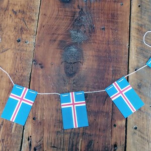 May include: A string of Icelandic flags, each featuring a blue background with a white cross and a red crossbar, hangs against a rustic wooden backdrop.