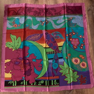Peut inclure: Un foulard carré en soie avec un motif floral vibrant. Le foulard présente une bordure rose et un motif coloré de fleurs, de feuilles et de formes abstraites dans des tons de violet, vert, bleu et orange. Le foulard est plié et posé sur une surface en bois.