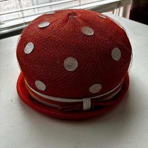 Puede incluir: Un sombrero redondo en forma de cúpula de color rojo anaranjado vibrante, con una fina red superpuesta adornada con pequeños detalles metálicos circulares de color plateado. La base está rodeada por una cinta blanca con un lazo.
